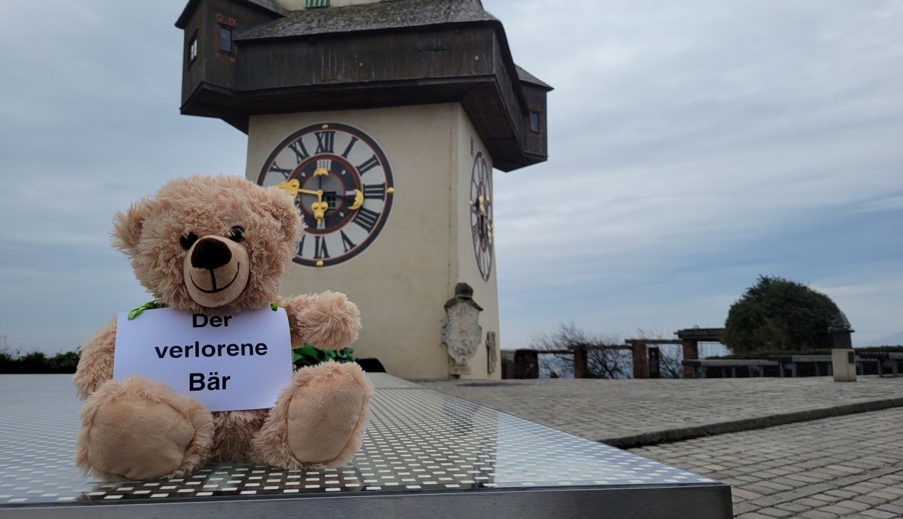 Ein Teddybär vor dem Grazer Schlossbild mit einem Schild, auf dem "Der verlorene Bär" steht.