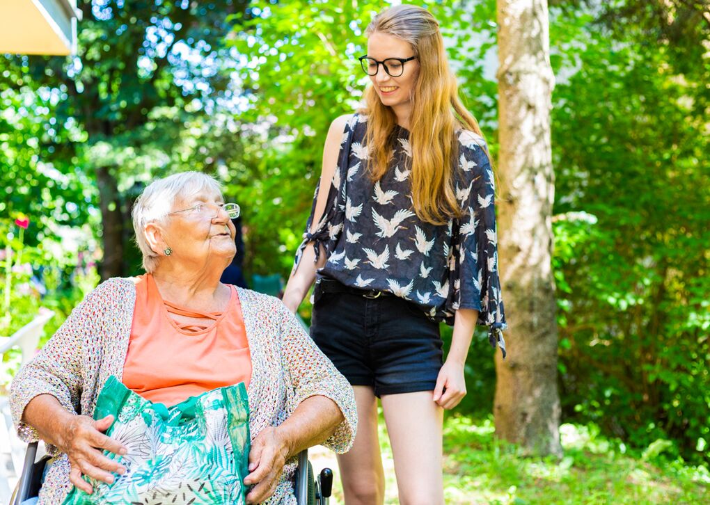 Eine junge Frau steht neben einer älteren Dame im Rollstuhl.