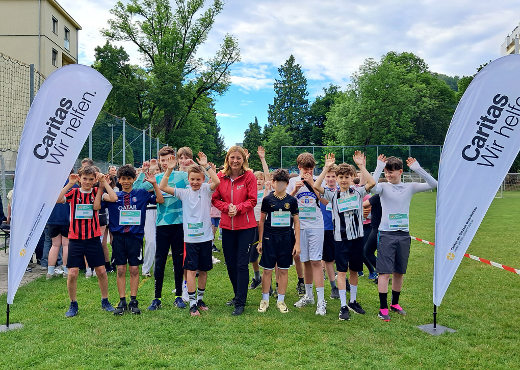 Schüler*innen in Laufkleidung heben gemeinsam mit Caritas-Direktorin Nora Tödtling-Musenbichler die Arme. Sie stehen auf einer Laufbahn, rechts und links von ihnen sind Flaggen mit dem Caritas-Logo.
