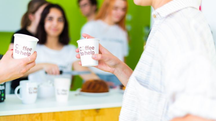 Zwei Personen stoßen mit einem Coffee to help-Becher an.