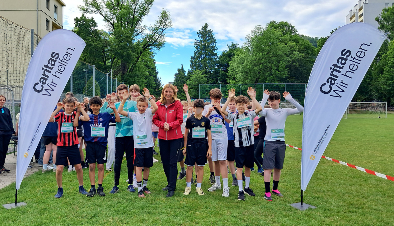Schüler*innen in Laufkleidung heben gemeinsam mit Caritas-Direktorin Nora Tödtling-Musenbichler die Arme. Sie stehen auf einer Laufbahn, rechts und links von ihnen sind Flaggen mit dem Caritas-Logo.