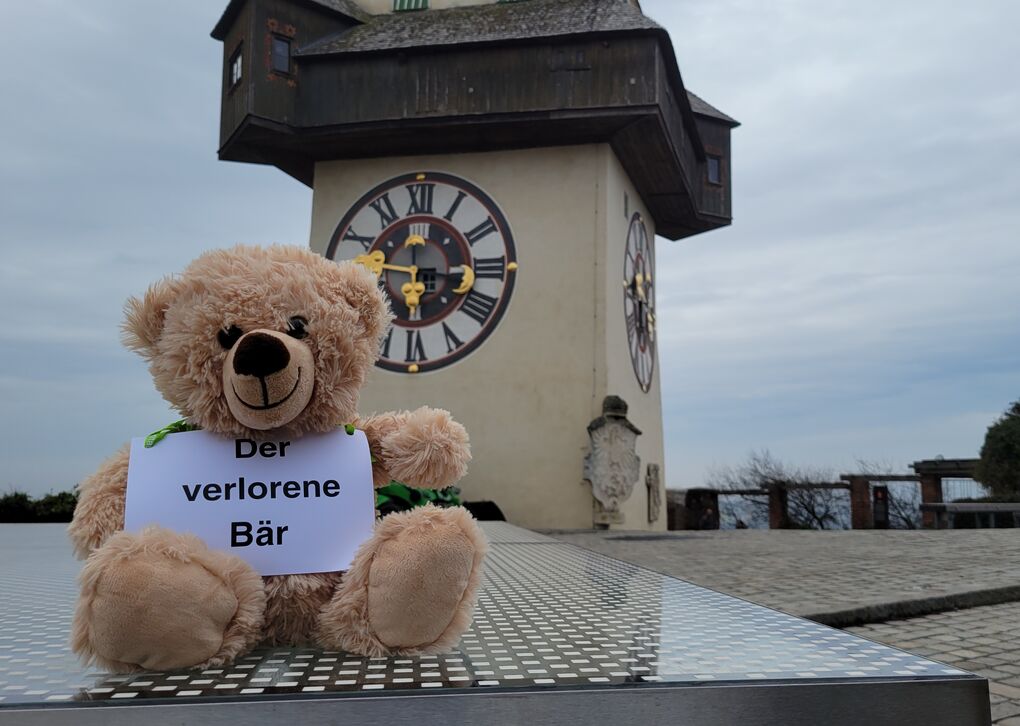Ein Teddybär vor dem Grazer Schlossbild mit einem Schild, auf dem "Der verlorene Bär" steht.