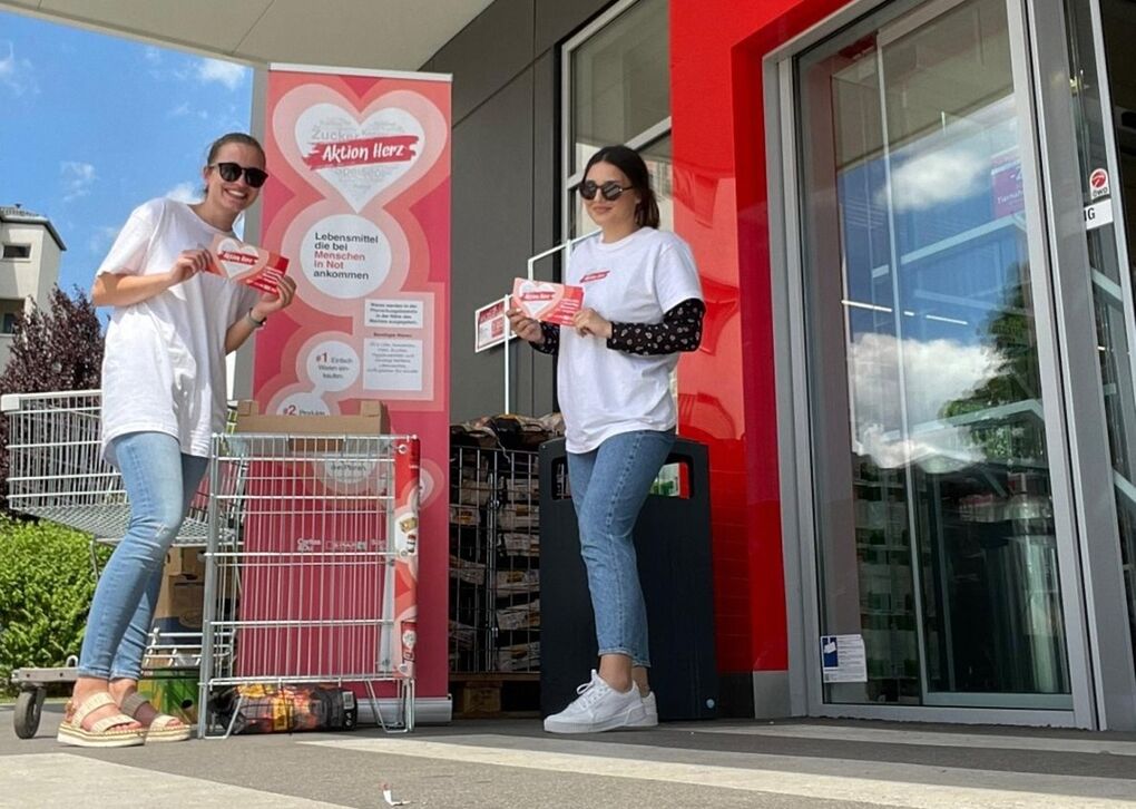 Zwei freiwillige Helferinnen stehen mit Aktion Herz T-Shirts, Flyern und Roll-Up vor einem SPAR Supermarkt.