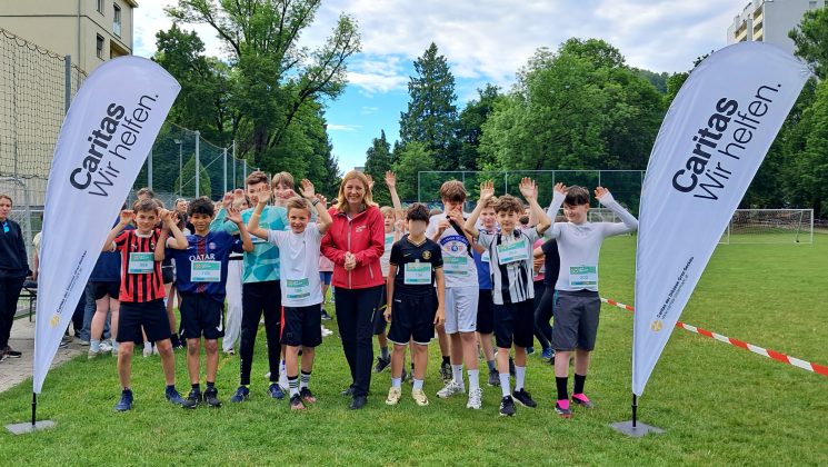 Schüler*innen in Laufkleidung heben gemeinsam mit Caritas-Direktorin Nora Tödtling-Musenbichler die Arme. Sie stehen auf einer Laufbahn, rechts und links von ihnen sind Flaggen mit dem Caritas-Logo.