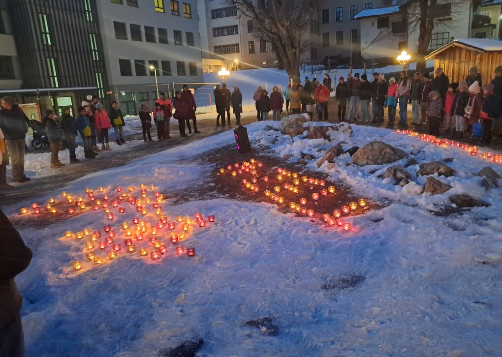 Menschen stehen vor brennenden Teelichtern im Schnee. Die Teelichter bilden Muster.