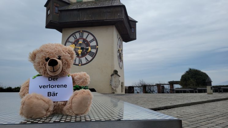 Ein Teddybär vor dem Grazer Schlossbild mit einem Schild, auf dem "Der verlorene Bär" steht.