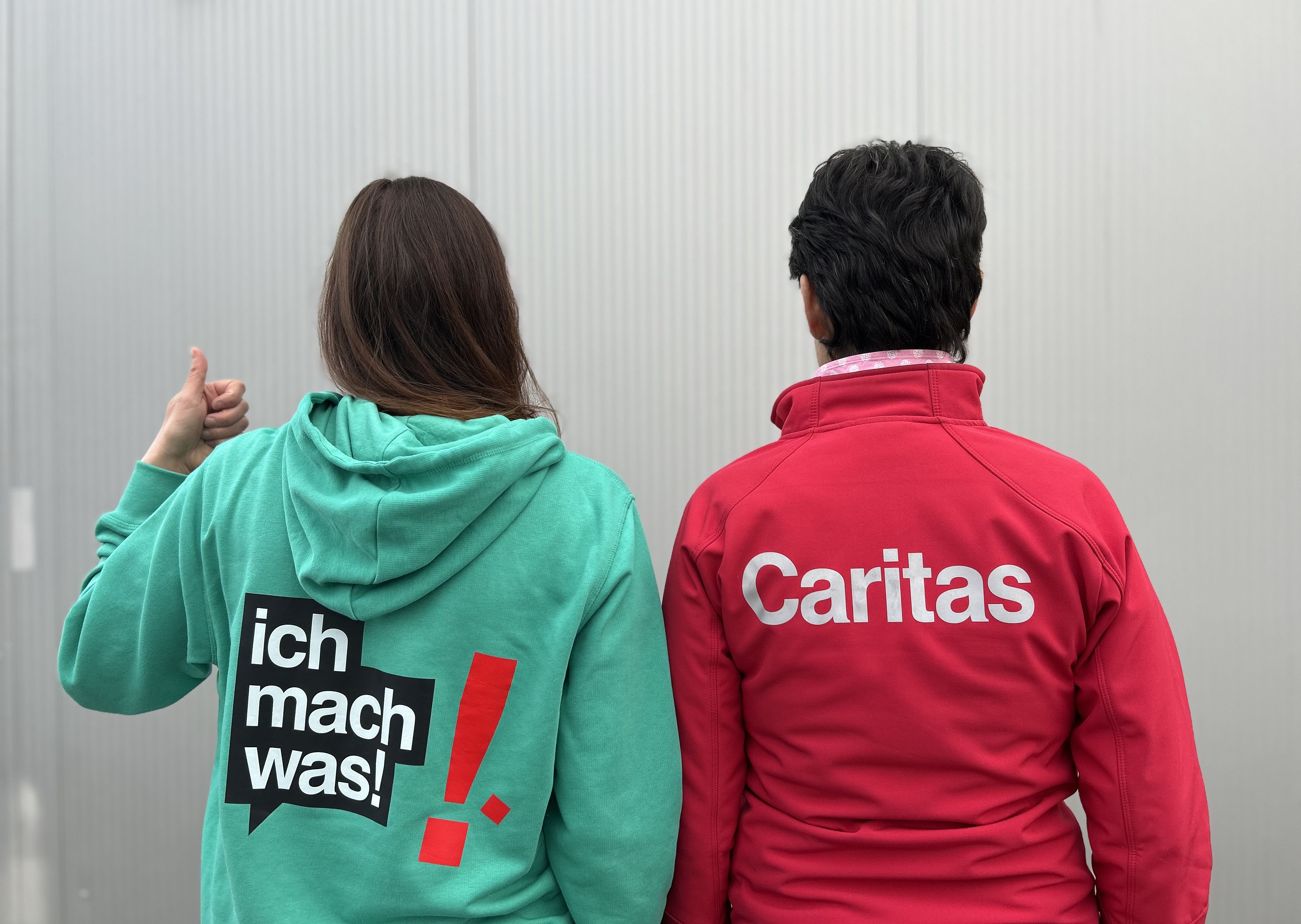 2 Frauen stehen mit dem Rücken zur Kamera. Auf einer Jacke steht "ich mach was" auf der anderen "Caritas".