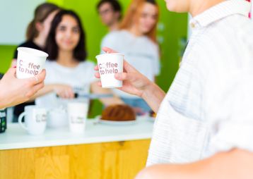 Zwei Personen stoßen mit einem Coffee to help-Becher an.