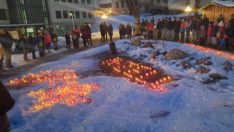 Menschen stehen vor brennenden Teelichtern im Schnee. Die Teelichter bilden Muster.
