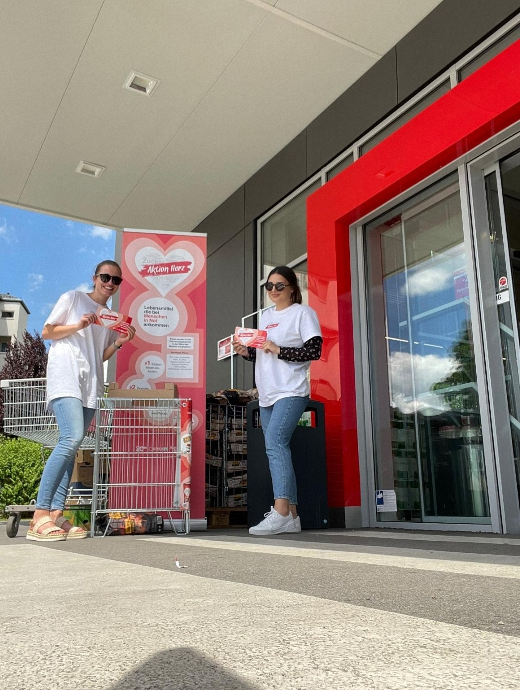 Zwei freiwillige Helferinnen stehen mit Aktion Herz T-Shirts, Flyern und Roll-Up vor einem SPAR Supermarkt.