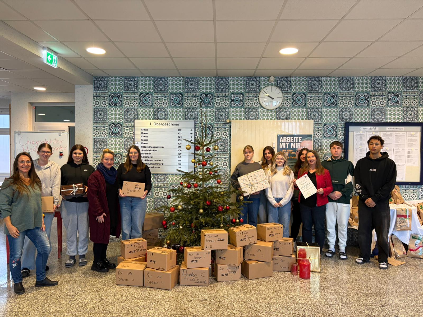 Schüler und Schülerinnen stehen neben einem Christbaum und halten Dankesurkunden in der Hand. Vor de Christbaum sind Kartons voller Sachspenden gestapelt.