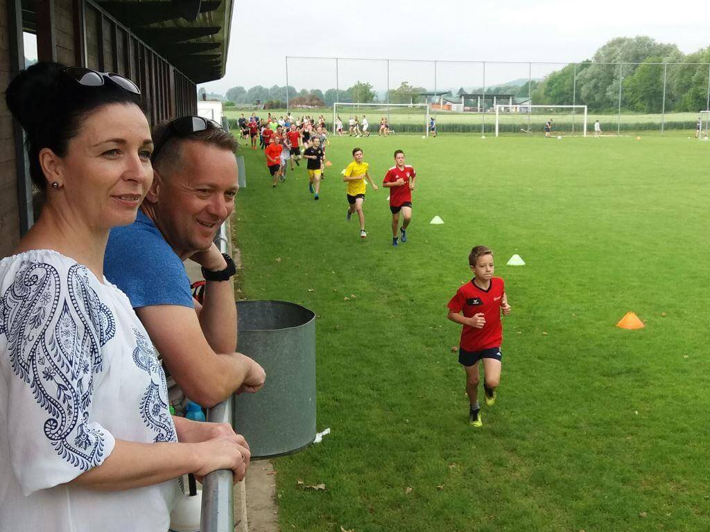 Zwei Erwachsene schauen Kinder beim Laufen auf dem Fußballplatz zu.
