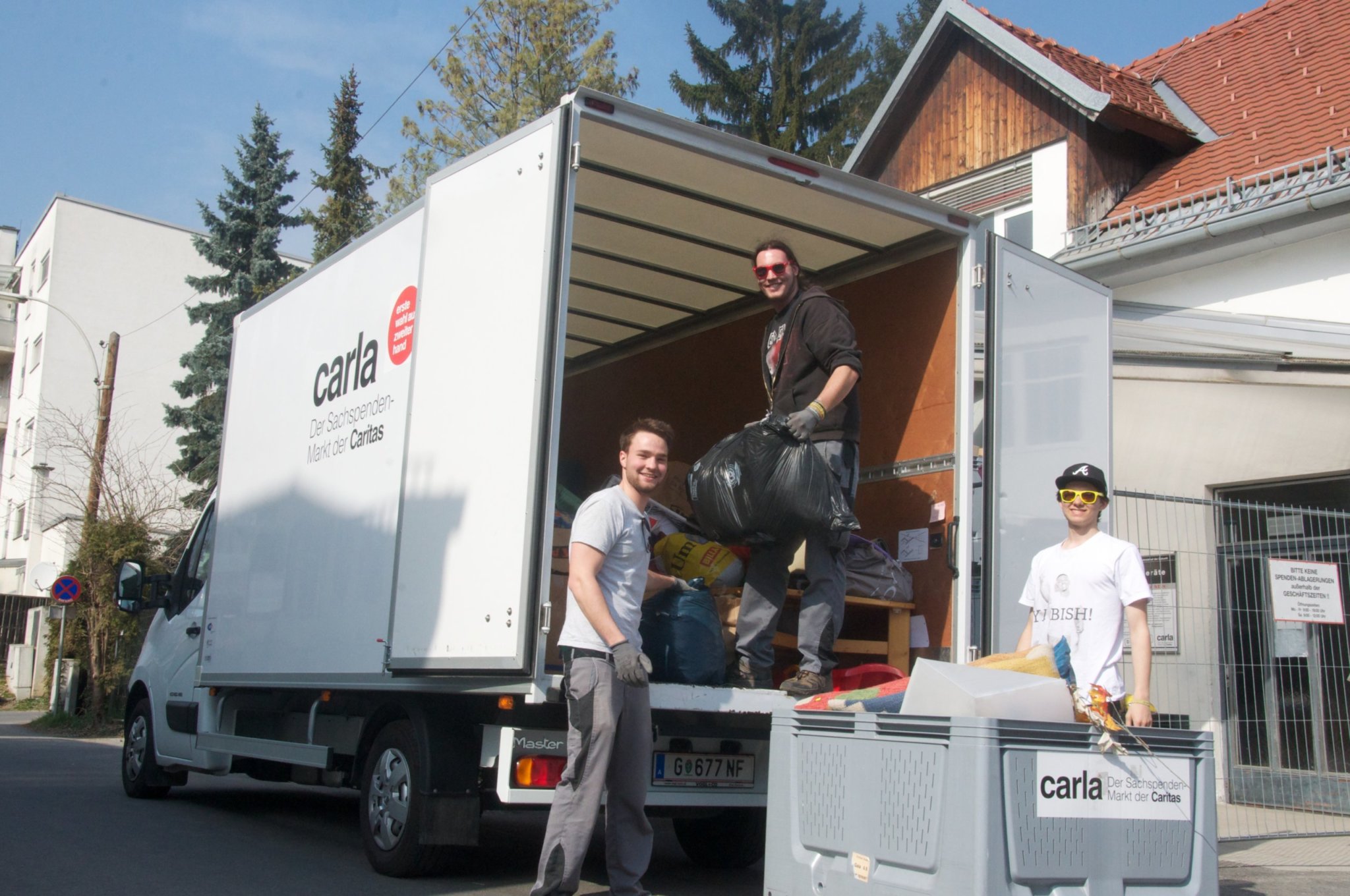 Drei junge Männer beim Beladen eines weißen Transportwagens mit dem Schriftzug 