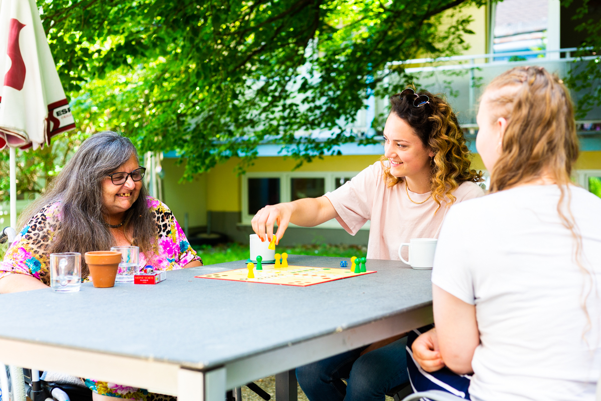 Zwei junge Frauen bei ihrem freiwilligen Engagement: Sie spielen Mensch ärgere dich nicht mit einer älteren Dame aus dem Pflegewohnhaus.